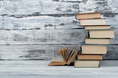 A stack of colorful books arranged against a rustic, whitewashed wooden background. One book is open, standing upright in front of the stack, creating a sense of curiosity and exploration. This image represents the pursuit of knowledge and the journey of discovery, similar to how Exposure Therapy Designs empowers individuals to uncover and understand their inner stories and patterns, fostering growth and healing.